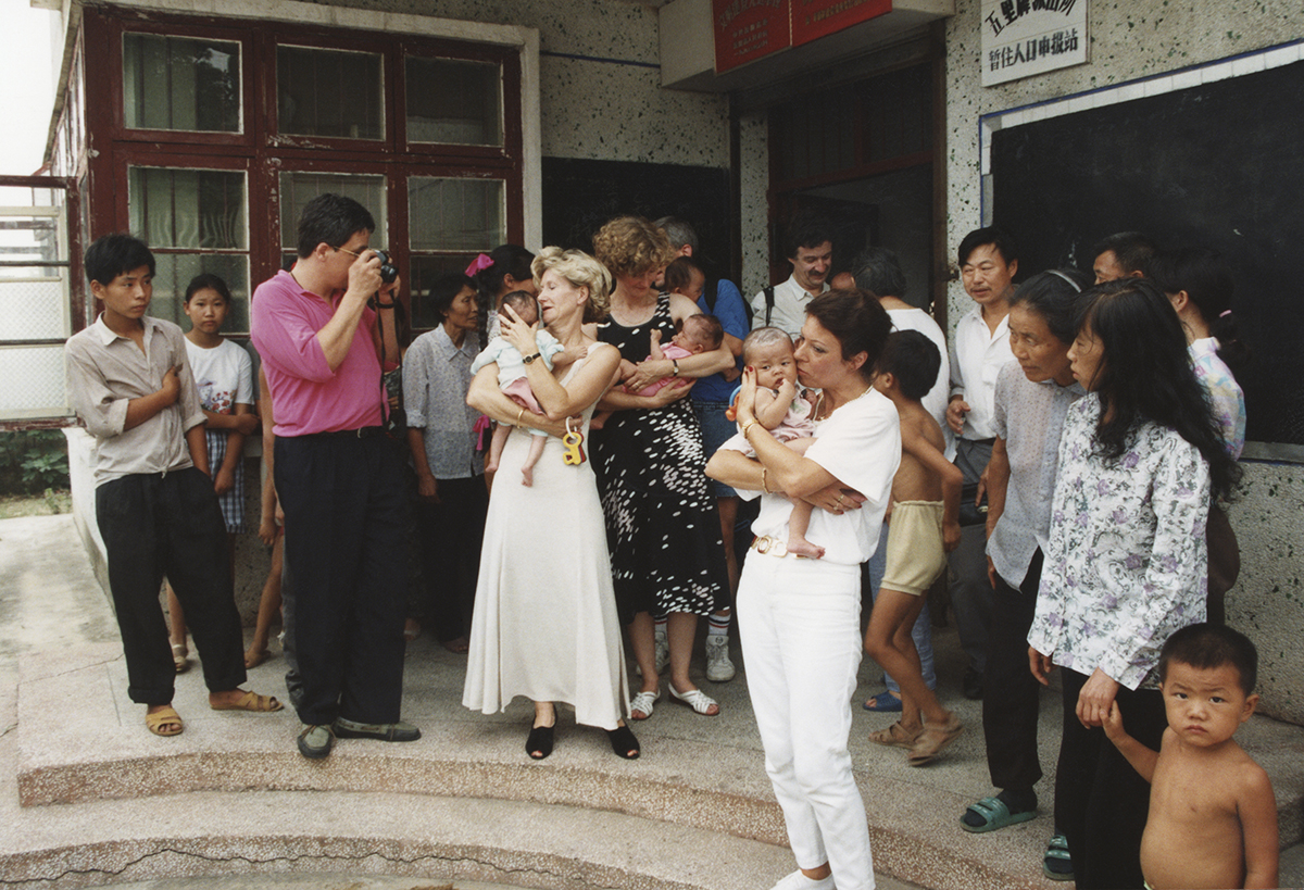 Mehrere Erwachsene stehen vor einem heruntergekommenen Gebäude und halten Babys und kleine Kinder auf dem Arm