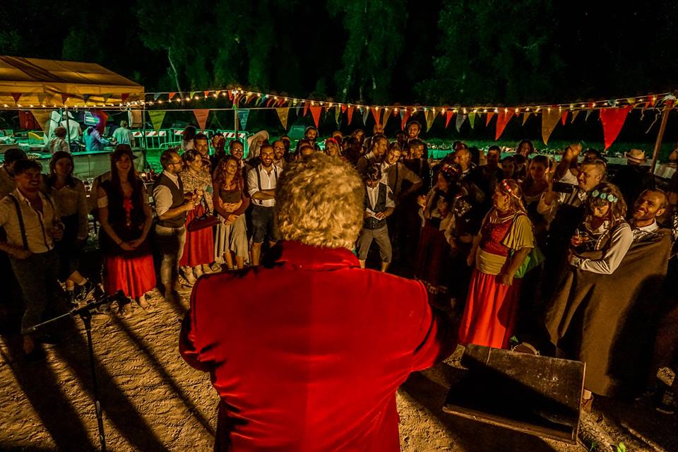 Ein Mann in roter Samtjacke und mit grauen Haaren steht mit dem Rücken zur Kamera und blickt auf versammelte Partygäste, die alle wie Hobbits gekleidet sind