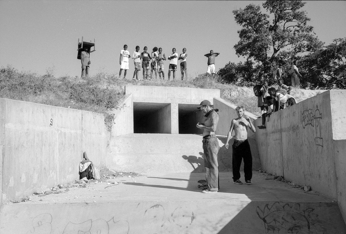 Africa, skateboarding, Jonas Camps - Black and white photo of a group of boys stood on a ridge looking down at men stood on a concrete floor.