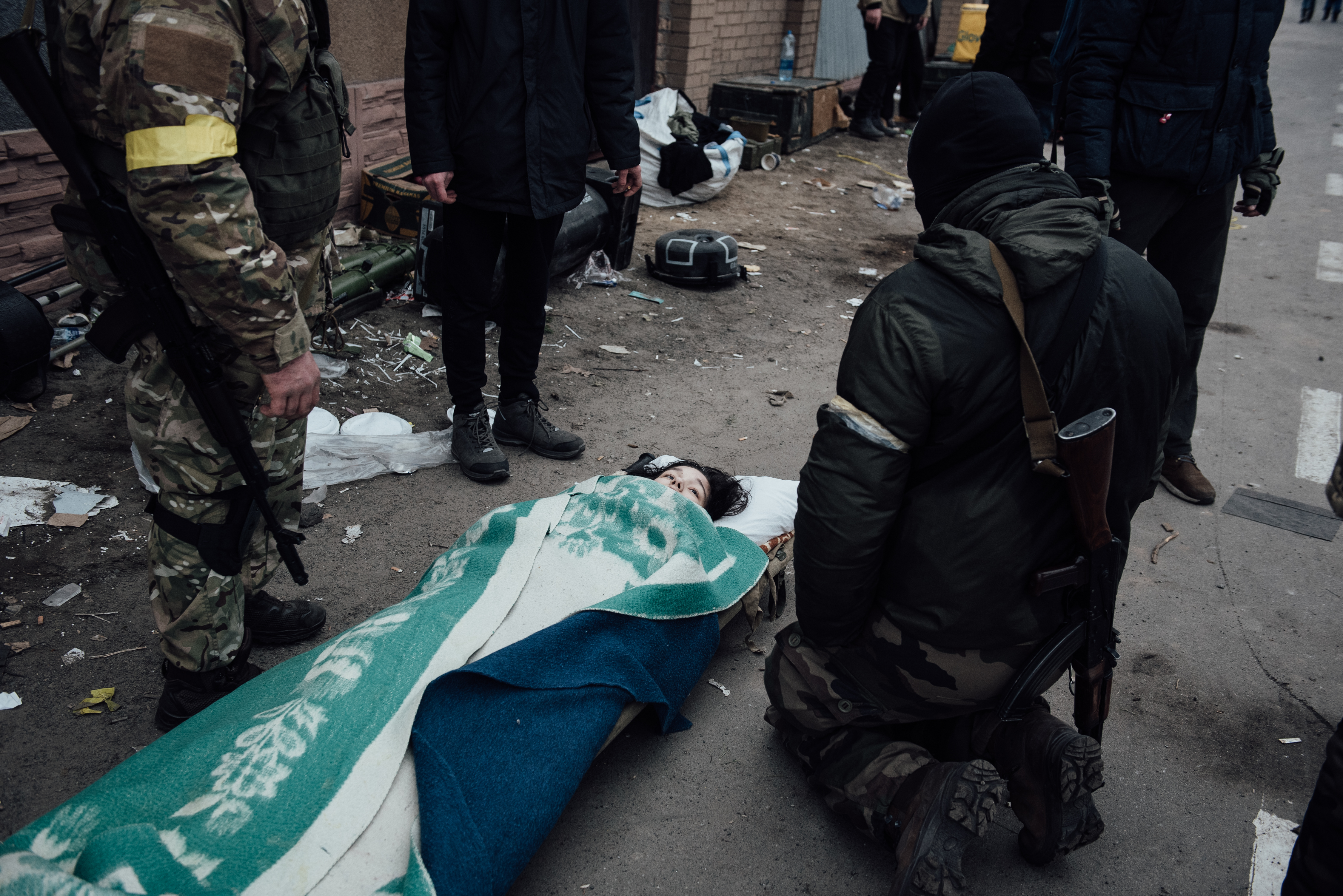 Adrien Vautier, Romanovka, Ukraine – young girl with black hair, lying on a stretcher on the ground, surrounded by soldiers.
