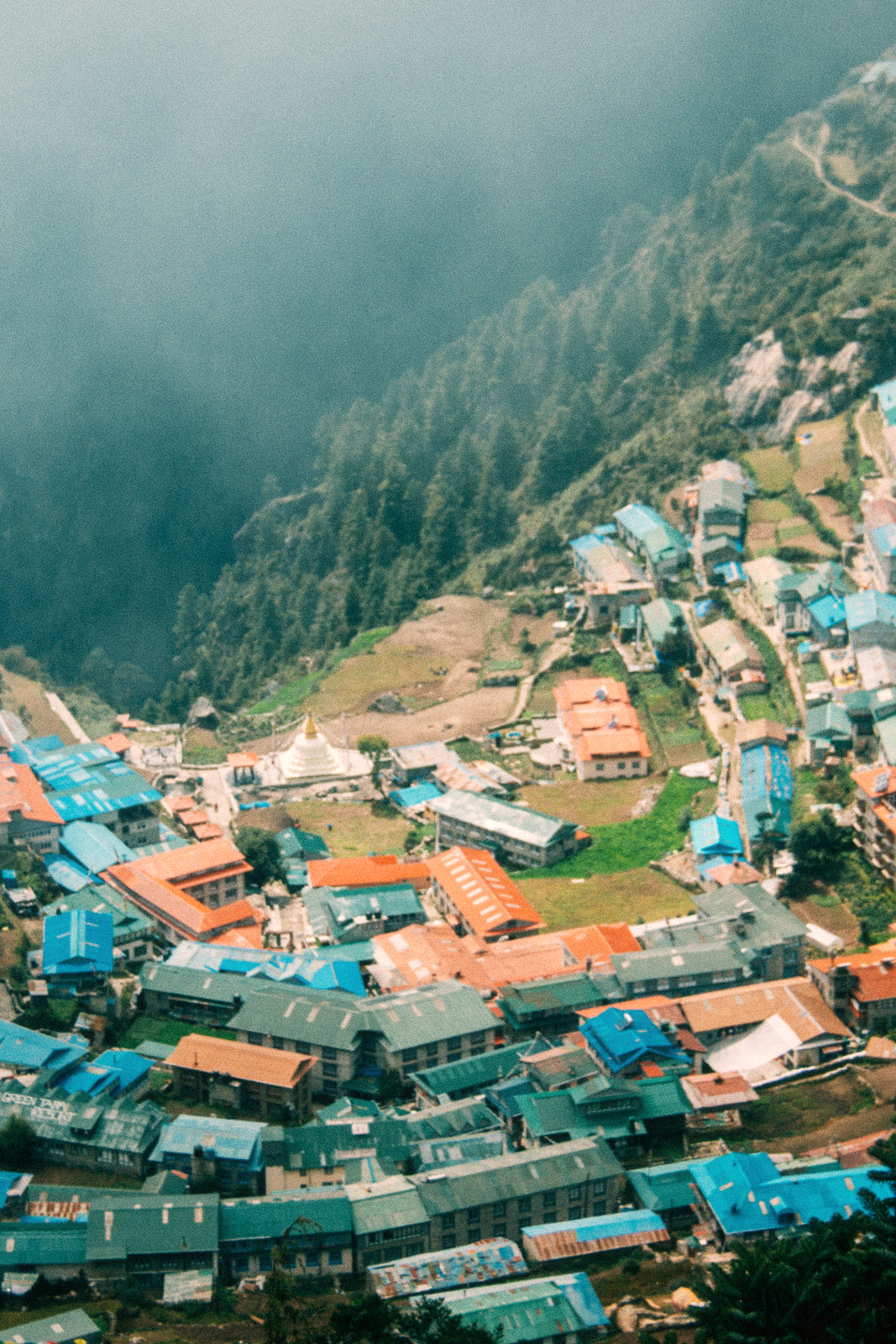 Namche bazar, Nepal – arial photo of a town perched near a steep cliff covered in pine forest. The houses' roofs are green and red and there's a small white and gold temple in the centre.