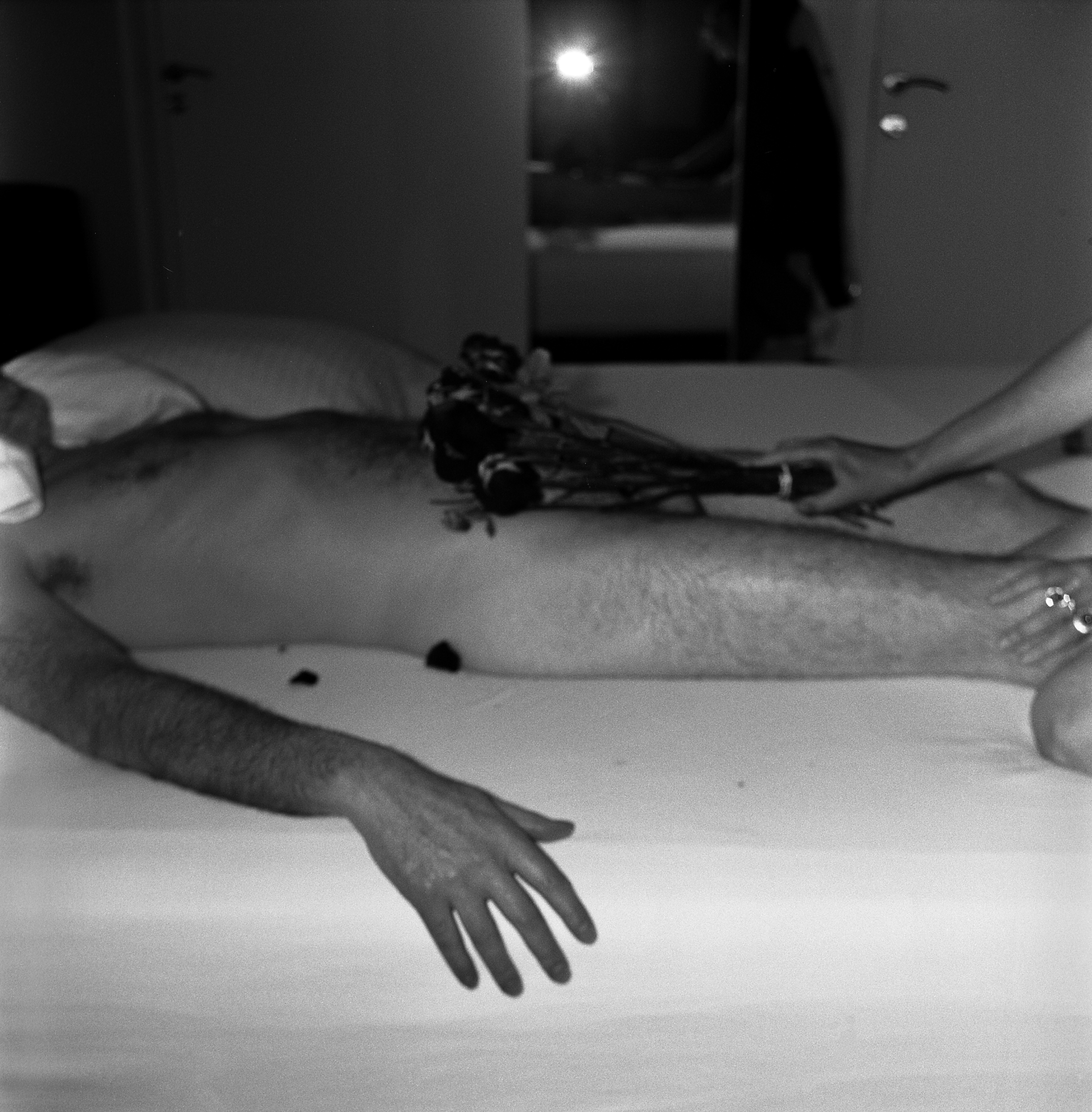 Photo of a hairy middle-aged man laying naked on a bed as woman (outside of the frame) caresses him with a bouquet of roses.